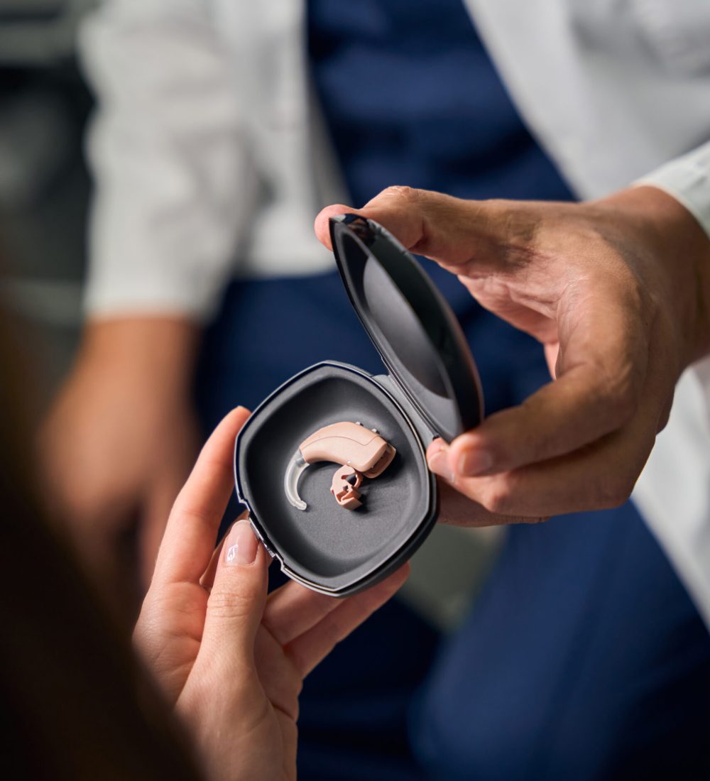 Elderly male doctor in a medical coat holding a black case with a hearing aid, showing it to a patient