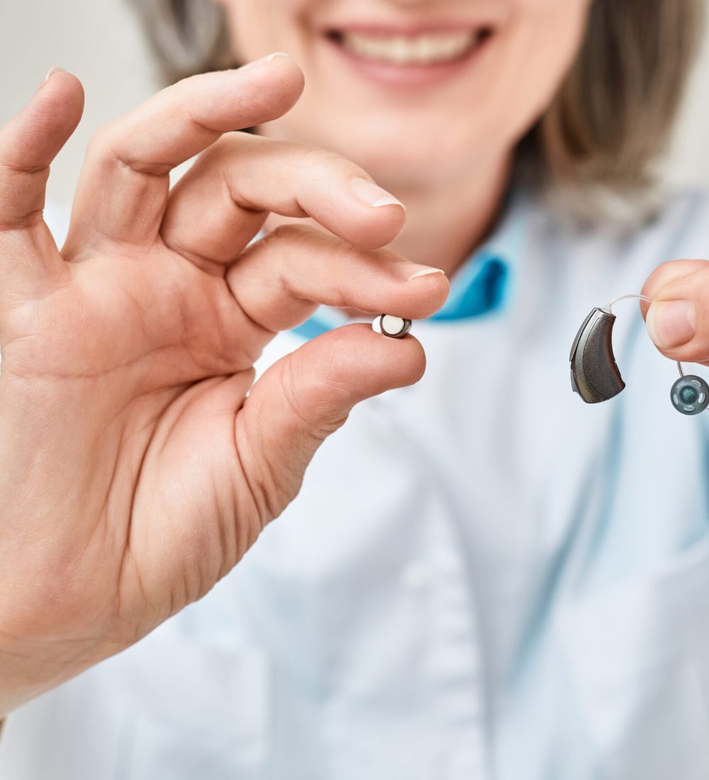 Hearing aid battery replacement. Positive audiologist showing hearing aid and battery for it, holding them in her hands in front of him