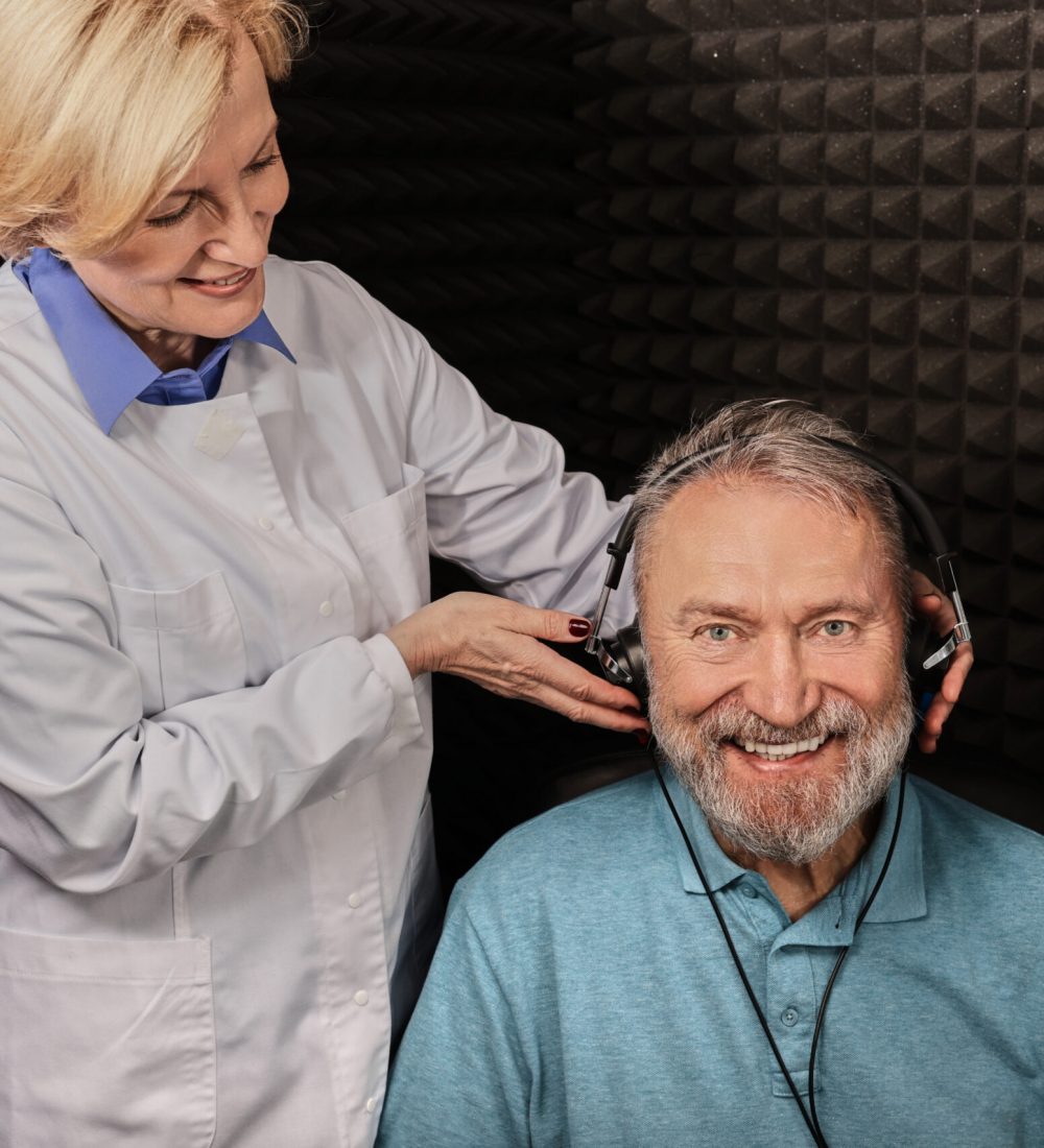 Portrait senior man while hearing check-up with audiologist at soundproof audiometric booth using audiometry headphones and audiometer